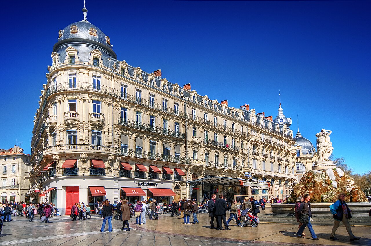 Place de la Comédie à Montpellier, immeuble à dôme, stores rouges, fontaine des Trois Grâces au premier plan.